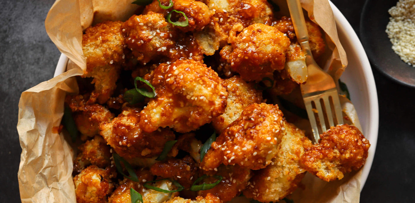 Sweet and savory Honey Garlic Cauliflower Poppers made with cauliflower, honey, and garlic, dredged in flour with Garlic Lemon Pepper seasoning, egg, breadcrumbs, seasoned with scallions and sesame seeds, served in a bowl on parchment paper with a fork.